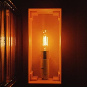 K&ouml;nnte beinhalten: Eine warme, leuchtende Gl&uuml;hbirne in einem quadratischen, orangefarbenen Rahmen. Die Gl&uuml;hbirne ist eine klare Gl&uuml;hbirne mit einem Filament aus Glas und einem silbernen Sockel.