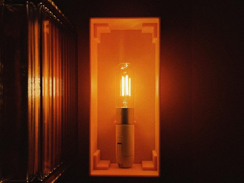 K&ouml;nnte beinhalten: Eine warme, leuchtende Gl&uuml;hbirne in einem quadratischen, orangefarbenen Rahmen. Die Gl&uuml;hbirne ist eine klare Gl&uuml;hbirne mit einem Filament aus Glas und einem silbernen Sockel.
