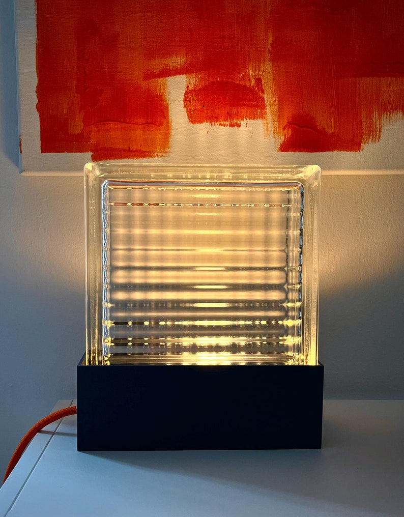 K&ouml;nnte beinhalten: Eine Glaslampe mit einem klaren, strukturierten Glasschirm und einem schwarzen Sockel. Die Lampe ist eingeschaltet und strahlt ein warmes Licht aus.