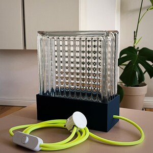 K&ouml;nnte beinhalten: Eine klare Glaslampe mit schwarzem Sockel und gelbem Kabel. Die Lampe hat ein strukturiertes Glasdesign und einen wei&szlig;en Schalter am Kabel.