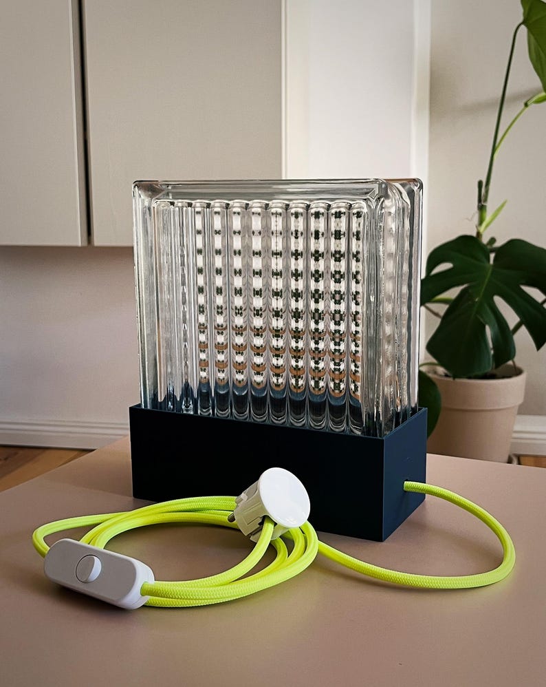 K&ouml;nnte beinhalten: Eine klare Glaslampe mit schwarzem Sockel und gelbem Kabel. Die Lampe hat ein strukturiertes Glasdesign und einen wei&szlig;en Schalter am Kabel.
