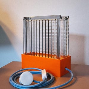K&ouml;nnte beinhalten: Eine Glaslampe mit gerippter Textur und einem orangefarbenen Sockel. Die Lampe hat ein blaues Kabel mit einem wei&szlig;en Stecker.