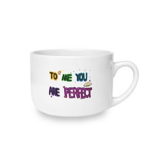 May include: A white ceramic mug featuring the colorful text TO ME YOU ARE PERFECT. The design includes a small orange star, a tiny planet with rings, and small heart accents. The mug has a smooth finish and a curved handle on the right side.