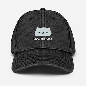 May include: A black baseball cap with a worn look. The cap displays a light blue cat face graphic and the white word "HAJIMAAA" on the front. It has a curved peak and a button on the crown, typical of this style of hat.