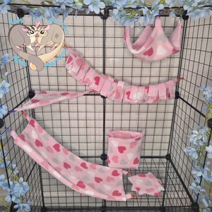 May include: A set of pink fleece pet accessories for a wire cage, including a hanging hammock, a fringed bridge, a corner hammock, a square pouch, and a small mat. Each item features a repeating pattern of pink, red, and white hearts.
