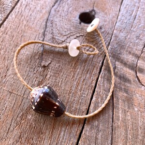May include: A handmade bracelet featuring a dark brown and white seashell pendant. The bracelet is strung on a natural-colored cord and includes a small, round, white button closure. The bracelet is displayed on a rustic wooden surface.