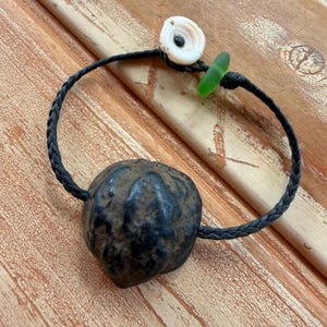 May include: A handmade bracelet featuring a dark, textured, round bead as a focal point. The bracelet includes a braided black cord, a white shell, and a small green glass piece. The bracelet is displayed on a wooden surface.