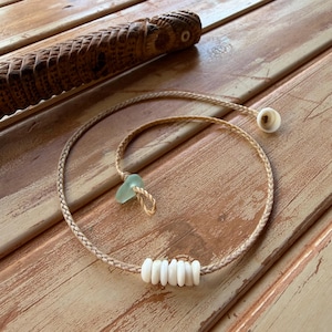 May include: Braided tan necklace with a light blue sea glass pendant and white shell beads. A carved wooden artifact is in the background. The necklace is displayed on a weathered wooden surface.