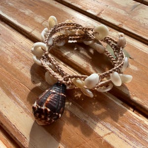 May include: A handmade bracelet featuring a woven, tan cord adorned with various white seashells. A large, brown and white seashell pendant hangs from the bracelet. The bracelet is displayed on a wooden surface.