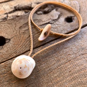May include: A handcrafted necklace featuring a large, cream-colored seashell pendant with a natural, braided cord. The necklace includes a smaller shell piece as a clasp. The seashell has a spiral pattern with light brown and pink hues.