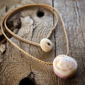 May include: A necklace with two shell pendants. The larger pendant is a pink and white spiral shell. The smaller pendant is a white shell with a brown center. The necklace is made of a braided brown cord.