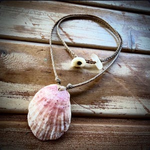 May include: A necklace featuring a pink and white scallop shell pendant suspended from a braided tan cord. The cord includes a small white shell bead and a translucent white piece of sea glass as a closure mechanism.