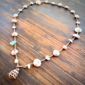 May include: A necklace featuring a tan braided cord strung with various small white seashells, a single smooth light green piece of sea glass, and a larger cone-shaped shell with a brown and white patterned exterior as the central pendant.