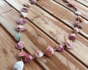 May include: A necklace featuring a series of small, variegated purple and white seashells strung along a tan cord. A single smooth, light green piece of sea glass is positioned on the left side, and a larger, textured white shell hangs as a pendant.