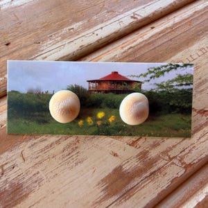 May include: A pair of beige, oval-shaped earrings with a textured surface. The earrings are displayed on a card featuring a landscape with a red-roofed building and green foliage.