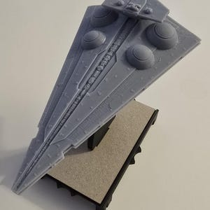 Puede incluir: Un modelo gris de un destructor estelar Star Destroyer impreso en 3D sobre una base gris. El modelo está detallado con una superficie texturizada y múltiples puertos de escape de motor.