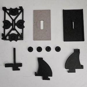 Puede incluir: Un conjunto de piezas de plástico negro para un modelo impreso en 3D. El conjunto incluye un marco con cuatro agujeros, dos piezas planas con un recorte rectangular, cuatro piezas pequeñas redondas, una pieza vertical con una pieza en forma de cruz unida y dos piezas angulares.