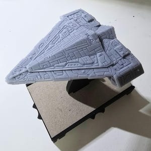 May include: A gray, 3D-printed model of a starship, featuring intricate details and a triangular design. The model is mounted on a black and tan base, showcasing the ship's complex structure. The ship's surface is covered in panels and geometric shapes.