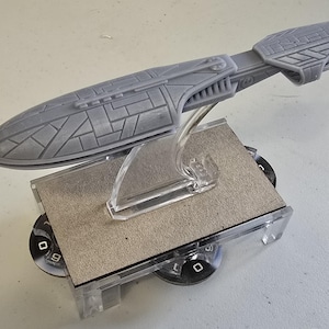Op de afbeelding: Een grijs ruimteschipmodel met gedetailleerde panelen, gemonteerd op een heldere plastic standaard. Het model staat op een rechthoekige basis met zwart-witte cirkelvormige elementen. Het ontwerp suggereert sciencefiction of fantasy.
