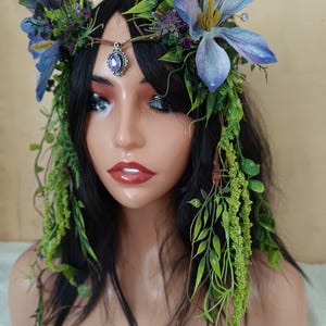 May include: A floral crown featuring large blue and purple flowers, green foliage, and a central purple jewel pendant. The crown is set against black hair and is designed to be worn on the head. The overall aesthetic is whimsical and natural.