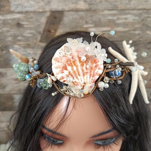 Może przedstawiać: Ozdoba do włosów w stylu syreny z dużą, pomarańczowo-białą muszlą jako centralnym elementem. Korona jest ozdobiona małymi białymi kwiatami, perłami, niebieskimi koralikami i rozgwiazdą. Podstawa to brązowa konstrukcja przypominająca gałązkę.