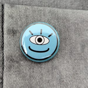 Può includere: Un bottone rotondo azzurro con un design a occhio sorridente. L'occhio è bianco con una pupilla nera e il bottone ha contorni neri e linee radianti. Il bottone è appuntato su un tessuto grigio.