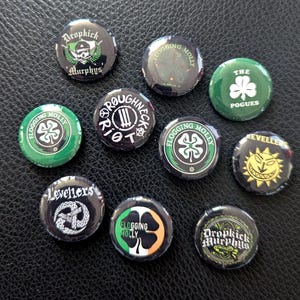 Könnte beinhalten: Zehn Kronkorken mit Bandlogos und Text, darunter Flogging Molly und Dropkick Murphys. Die Kronkorken zeigen Designs mit Kleeblättern, Totenköpfen und Sonnen in Grün, Schwarz und Weiß. Es werden verschiedene Schriftarten und Stile verwendet.