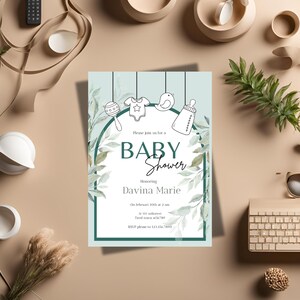 Op de afbeelding: Een lichtblauwe babyborrel uitnodiging met groene bladeren en een witte boog. De uitnodiging toont een babyratel, een rompertje, een vogel en een fles. De tekst luidt "U bent van harte welkom op onze babyborrel ter ere van Davina Marie Op 10 februari om 14:00 uur In 101 Milltown Tired Town 56789 RSVP graag naar 123 456 7890"