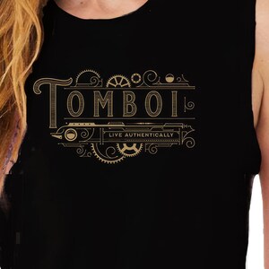 May include: Black sleeveless tank top with the word "TOMBOI" in gold lettering, featuring a decorative design with gears and the phrase "LIVE AUTHENTICALLY". The top is made of a soft, comfortable material.