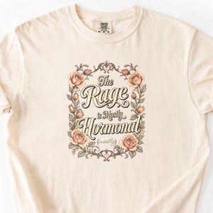 Op de afbeelding: Crèmekleurig T-shirt met de tekst "The Rage is Mostly Hormonal (mostly)" in een decoratieve bloemenframe. Het ontwerp bevat perzikrozen en groene bladeren. Het shirt heeft korte mouwen en een ronde hals.