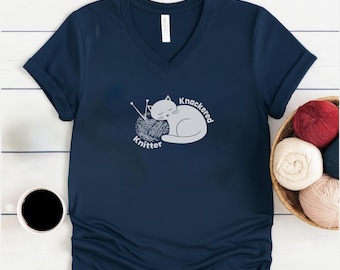 Le motif graphique Knackered Knitter avec un chat dormant sur une pelote de laine imprimé sur un t-shirt souple à col en V.