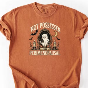 Op de afbeelding: Verbrand oranje t-shirt met de tekst "NOT POSSESSED, JUST PERIMENOPAUSAL". Het shirt heeft een afbeelding van een geest die een waaier in een spiegel vasthoudt, met vleermuizen, kaarsen en een halve maan.