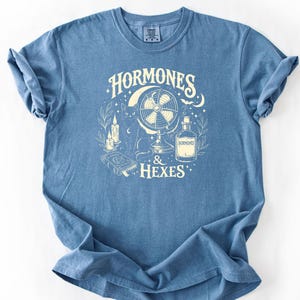 Op de afbeelding: Een blauw t-shirt met korte mouwen en een crèmekleurige grafische print van een kat, een vintage ventilator, kaarsen, een boek met een pentagram en een fles met het label HORMONES. De tekst HORMONES & HEXES staat in een decoratief lettertype.