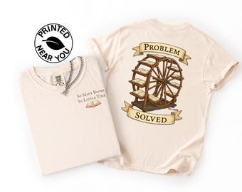 Könnte beinhalten: Zwei cremefarbene T-Shirts. Ein Shirt hat den Text "So Many Books So Little Time" und eine kleine Buchgrafik. Das andere Shirt zeigt ein Bücherkarussell mit dem Text "Problem Solved". Ein "Printed Near You"-Logo ist sichtbar.