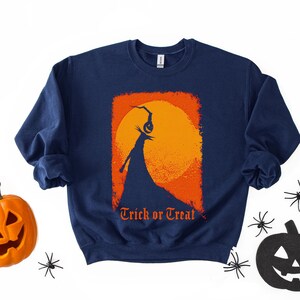 Puede incluir: Sudadera azul marino con un dise&ntilde;o tem&aacute;tico de Halloween. El gr&aacute;fico presenta una silueta de un personaje sosteniendo una linterna de calabaza contra una luna naranja, enmarcada por un rect&aacute;ngulo naranja. Las palabras "Trick or Treat" est&aacute;n impresas debajo.