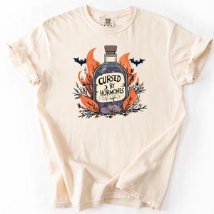 Op de afbeelding: Crèmekleurig T-shirt met een afbeelding van een drankfles met de tekst "Cursed by Hormones". De fles is omgeven door oranje vlammen, bladeren en vleermuizen, wat een griezelig Halloween-thema creëert.