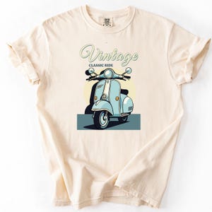 Op de afbeelding: Een crèmekleurig T-shirt met korte mouwen en een afbeelding van een lichtblauwe vintage scooter. De tekst "Vintage" staat in een cursief lettertype boven de scooter, met "CLASSIC RIDE" in een kleiner, vet, schreefloos lettertype eronder.