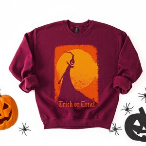Puede incluir: Sudadera burdeos de cuello redondo con un dise&ntilde;o gr&aacute;fico de tem&aacute;tica de Halloween. El dise&ntilde;o presenta una silueta de una figura sosteniendo una linterna de calabaza contra una luna naranja y amarilla, con las palabras "Trick or Treat" debajo. Una calabaza y ara&ntilde;as est&aacute;n a los lados.