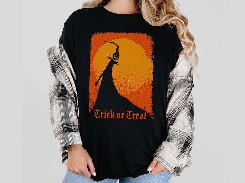 Puede incluir: Camiseta negra con un dise&ntilde;o gr&aacute;fico naranja y amarillo de una figura de Halloween contra una luna llena. Las palabras "Trick or Treat" est&aacute;n impresas en naranja debajo. Parte de una colecci&oacute;n tem&aacute;tica de Halloween.
