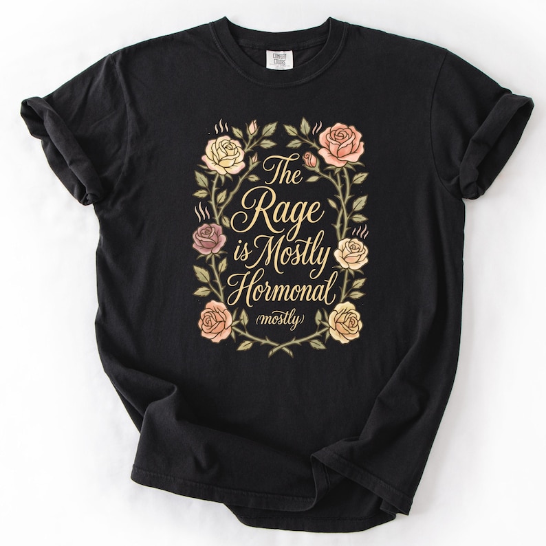 May include: Black t-shirt featuring the text "The Rage is Mostly Hormonal (mostly)" within a floral wreath. The design includes roses in pink, peach, and yellow. The shirt is made of a soft material.