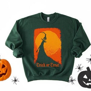 Puede incluir: Sudadera verde oscuro con un dise&ntilde;o tem&aacute;tico de Halloween. El dise&ntilde;o presenta una silueta de una figura sosteniendo una linterna de calabaza contra una luna naranja y amarilla, con las palabras "Trick or Treat" en naranja.