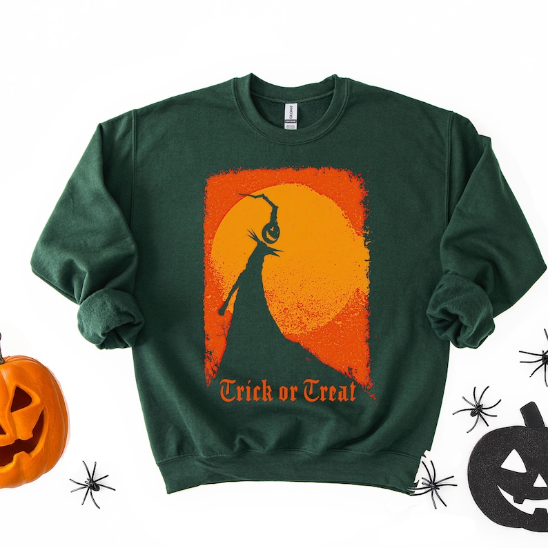 Puede incluir: Sudadera verde oscuro con un dise&ntilde;o tem&aacute;tico de Halloween. El dise&ntilde;o presenta una silueta de una figura sosteniendo una linterna de calabaza contra una luna naranja y amarilla, con las palabras "Trick or Treat" en naranja.
