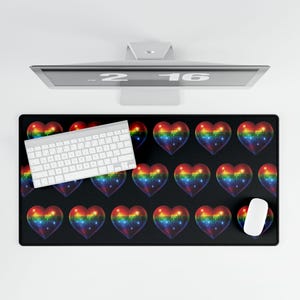 May include: Black rectangular desk mat featuring a pattern of rainbow-colored heart shapes. A white keyboard and mouse are on the mat. A computer monitor is visible in the background.