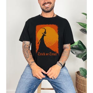 Puede incluir: Camiseta negra con un dise&ntilde;o tem&aacute;tico de Halloween. El dise&ntilde;o presenta la silueta de una figura con cabeza de calabaza contra un sol naranja, con las palabras "Trick or Treat" en naranja.