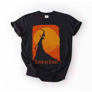 Puede incluir: Camiseta negra con un dise&ntilde;o de Halloween. El gr&aacute;fico incluye una figura silueteada con una cabeza de calabaza contra un sol naranja, con las palabras "Trick or Treat" en escritura naranja.