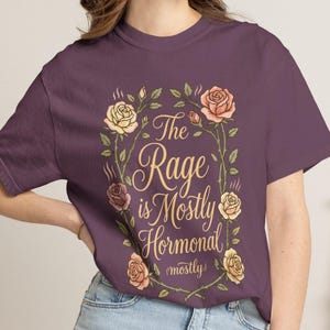 Może przedstawiać: Ciemnofioletowy t-shirt z centralną grafiką przedstawiającą wieniec z r&oacute;żowych i ż&oacute;łtych r&oacute;ż. Wewnątrz wieńca znajduje się napis "The Rage is Mostly Hormonal (mostly)" wykonany ozdobną, kursywną czcionką w kolorze złotym.