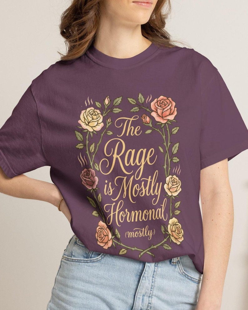 Może przedstawiać: Ciemnofioletowy t-shirt z centralną grafiką przedstawiającą wieniec z r&oacute;żowych i ż&oacute;łtych r&oacute;ż. Wewnątrz wieńca znajduje się napis "The Rage is Mostly Hormonal (mostly)" wykonany ozdobną, kursywną czcionką w kolorze złotym.