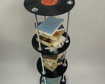 May include: A black vinyl record shelf with four tiers. The shelf is made of four black vinyl records with metal rods connecting them. The shelf is filled with books.