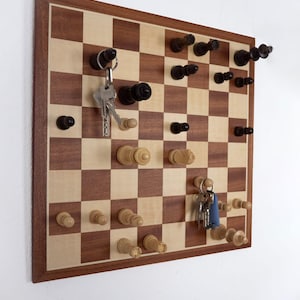 Könnte beinhalten: Ein hölzerner Schachbrett-Schlüsselhalter mit einem braunen und beigefarbenen Karomuster. Der Schlüsselhalter verfügt über Schachfiguren-Stifte zum Aufhängen von Schlüsseln. Mehrere Schlüsselsets hängen an den Stiften. Das Brett ist mit einem dunkelbraunen Rahmen versehen.