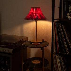 May include: A red and black patterned lampshade sits atop a brass lamp stand. The lamp is on a two-tiered side table made from vinyl records. A record player and a shelf of vinyl records are in the background.